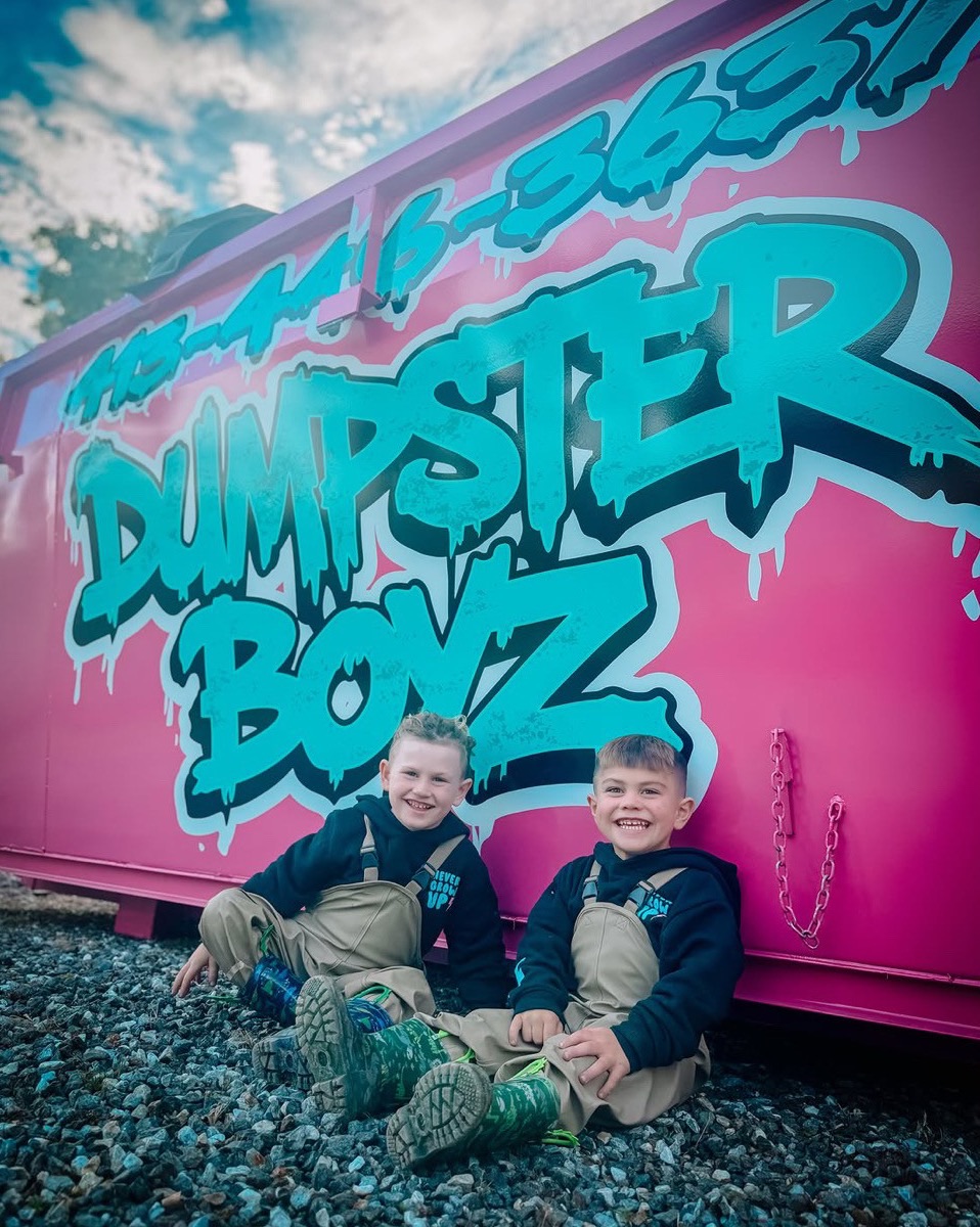 Two young brothers sitting on the gravel and grinning in front of a hot-pink Dumpster Boyz dumpster.