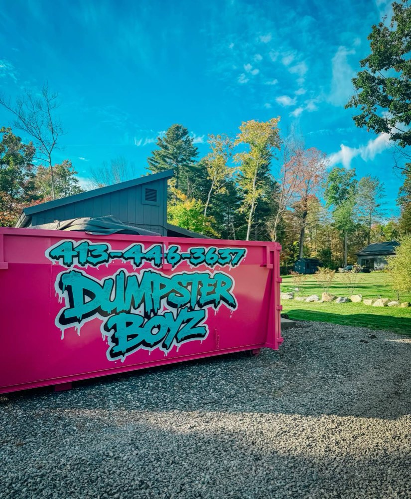 The hot-pink Dumpster Boyz dumpster with its teal logo and phone number, parked on a gravel driveway under a bright blue sky.